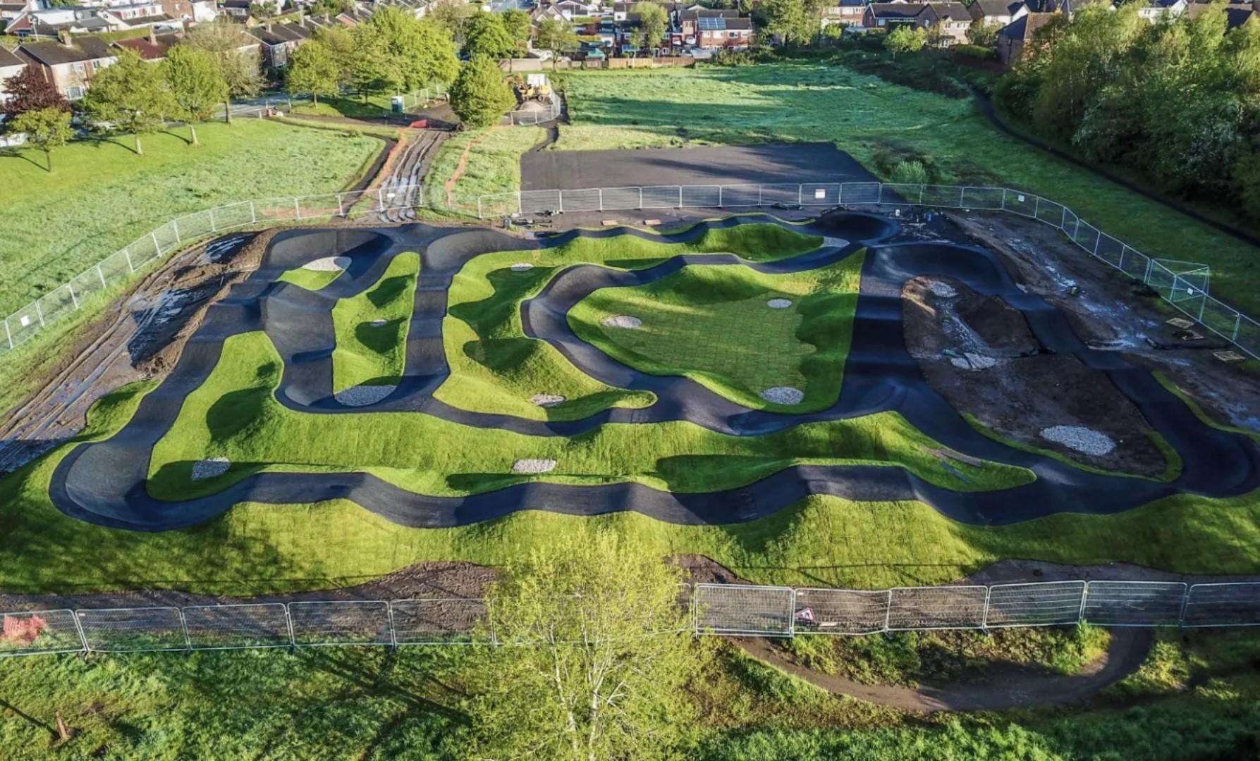 Aerial view of the Kidsgrove Pump Track, showing a winding network of smooth black asphalt paths and raised grassy mounds. The track is enclosed by temporary fencing and surrounded by open green fields, trees, and nearby houses in the background.