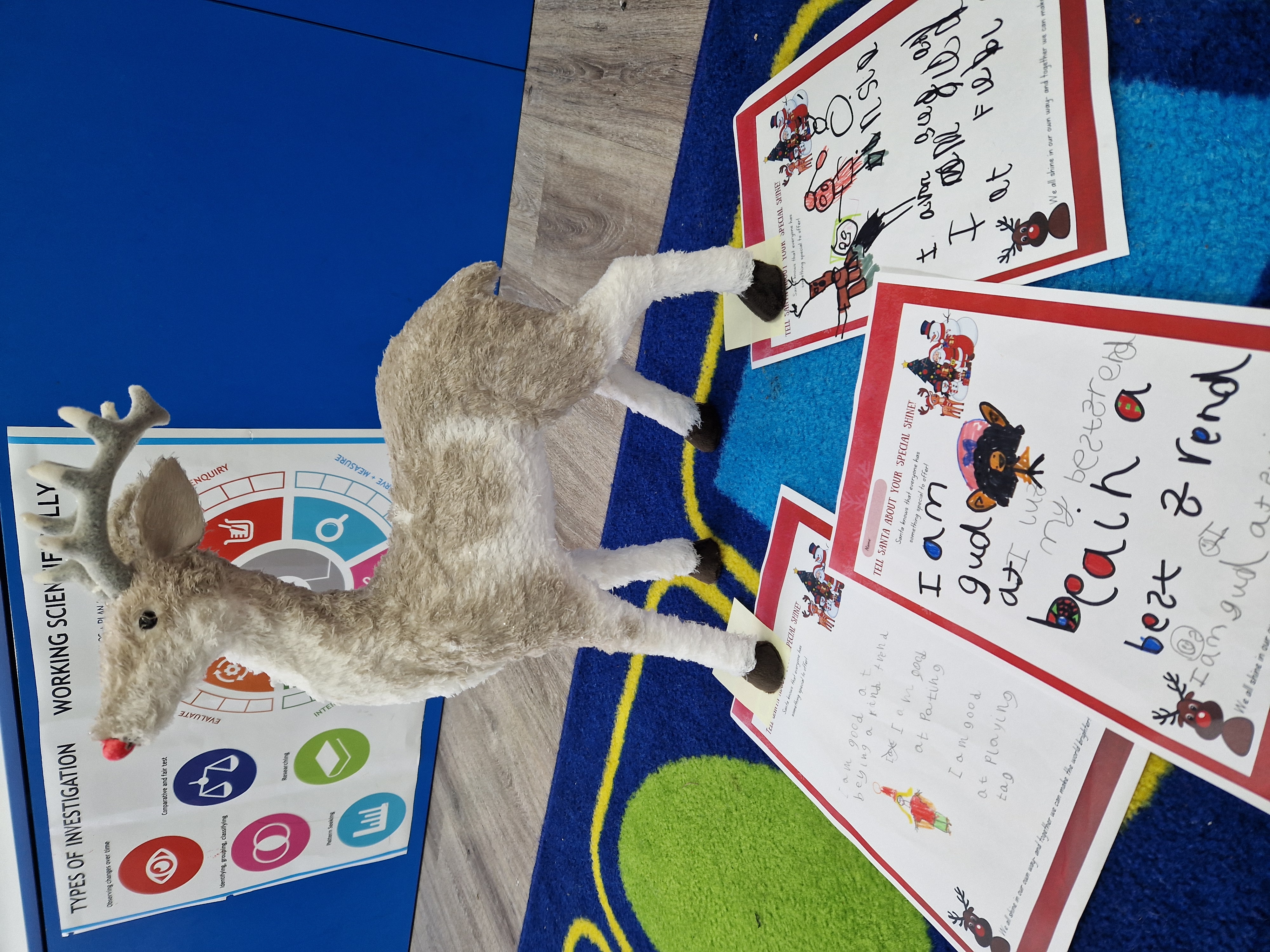 Photograph of a stuffed toy reindeer standing on a classroom rug. In front of him are examples of children's writing about inclusion, the front piece of work reads 'I am good at being a best friend.'