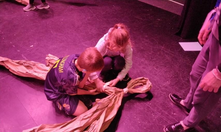 Colour photo of two children crouching down on a stage creating a stage prop out of brown paper.