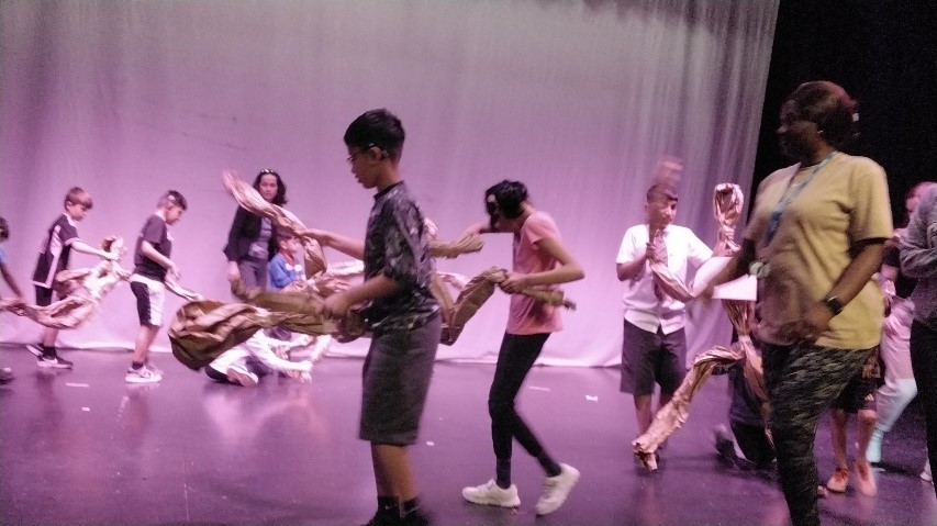 Photo shows a group of children walking round in a circle on a stage, holding props made from brown paper.