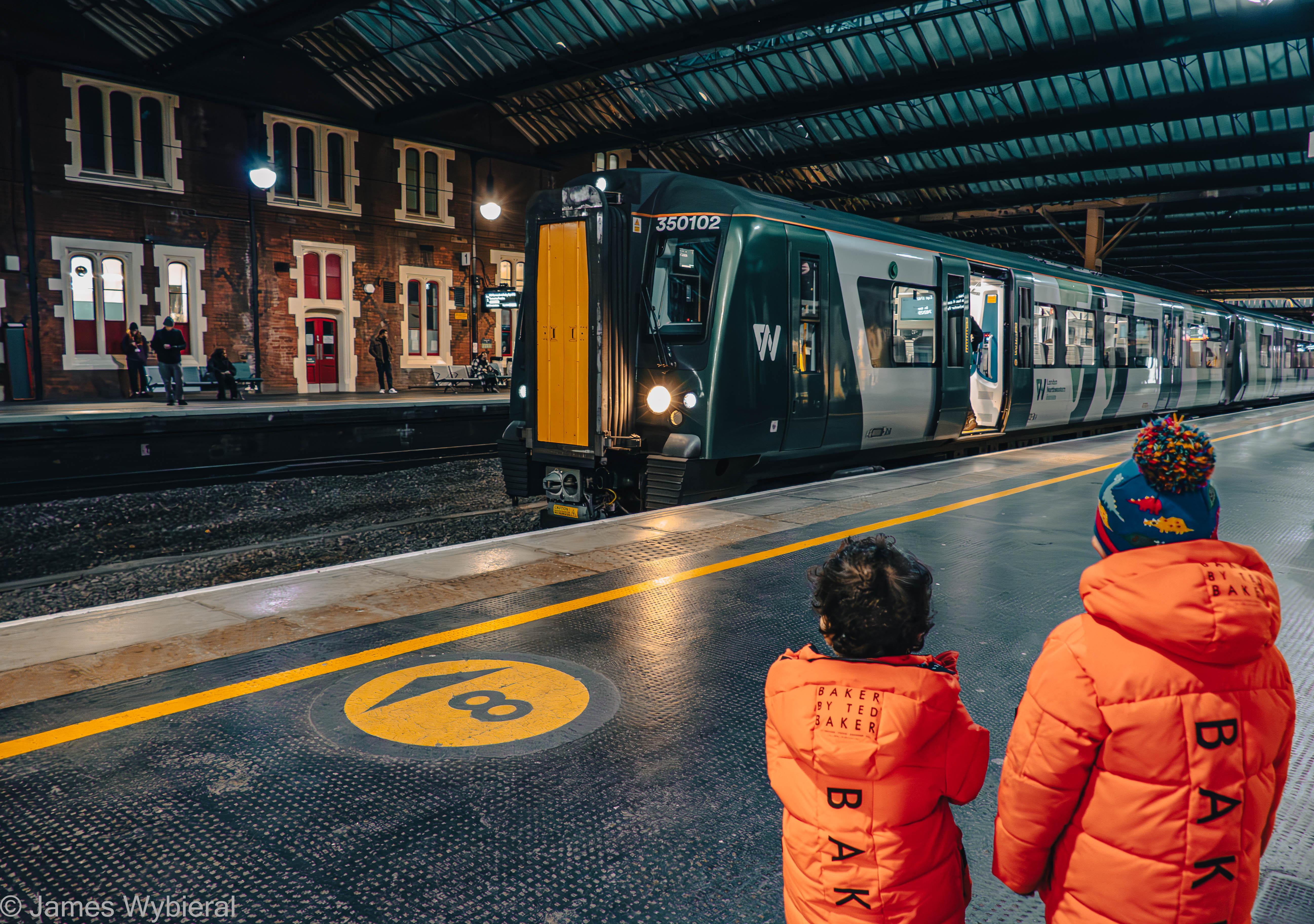 Photograph showing the back of two small children in orange winter coats standing on the platform of a railway station watching a train pull in.