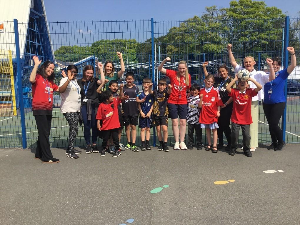 Photograph showing group of primary age children, teaching staff and Debbie Patterson standing on a playground. All are wearing sports clothes and are smiling and waving.