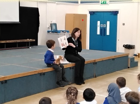 A teacher and pupil at the front of a school hall with the rest of the children sitting facing them,  in a signed storytelling session, using props and visual communication to bring the story to life.